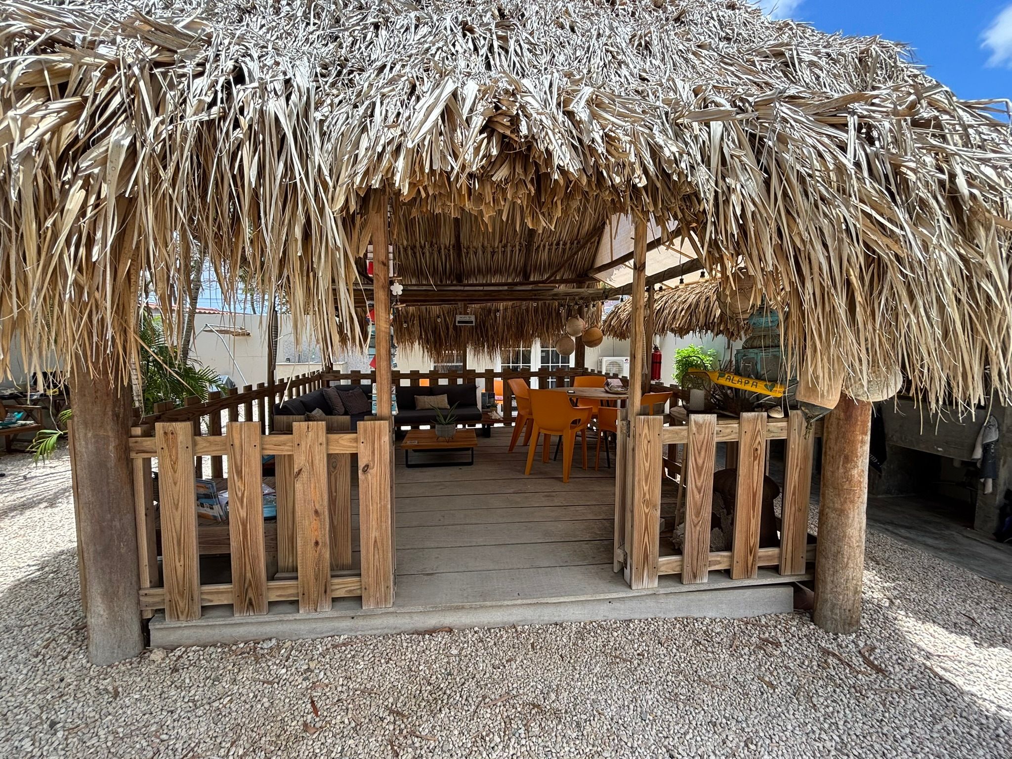 A thatched-roof outdoor seating area with wooden furniture and a gravel floor.
