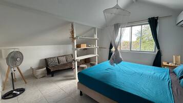 A bedroom with a bed, a standing fan, a sofa, and a window with curtains.