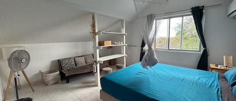 A bedroom with a bed, a standing fan, a sofa, and a window with curtains.
