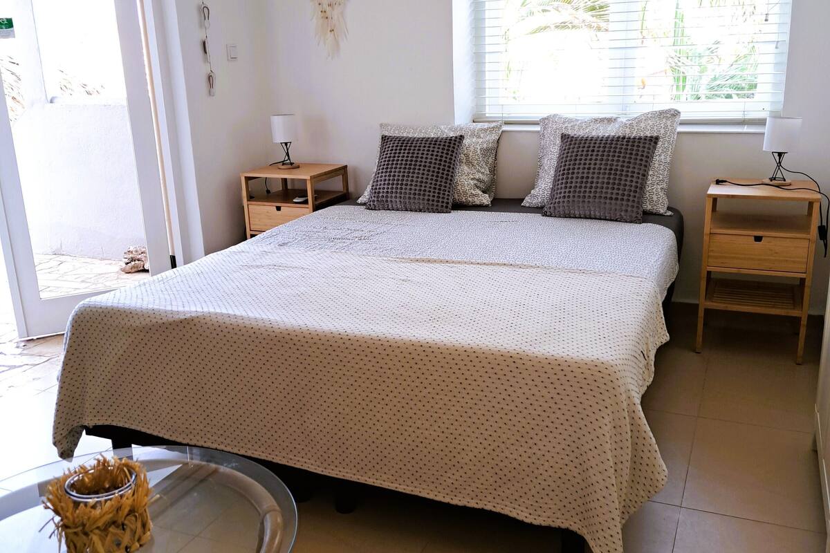 Une chambre à coucher avec un grand lit, deux tables de chevet, une fenêtre avec des stores et une table en verre ornée d’un objet décoratif.