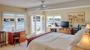 Habitación de hotel con cama, escritorio, silla, televisor, chimenea y ventanas amplias con vistas a una masa de agua y casas.