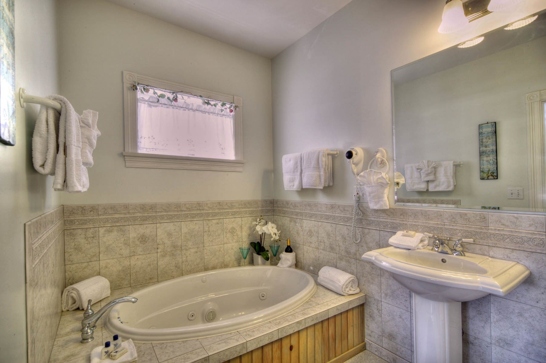 A bathroom with a bathtub, a sink, and a large mirror. There are towels hanging on the wall and a window with a curtain.