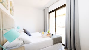 Una cama bien tendida con sábanas blancas, una almohada gris, una almohada verde azulada y una cesta con un croissant, un vaso de zumo de naranja y un pequeño jarrón con una flor.