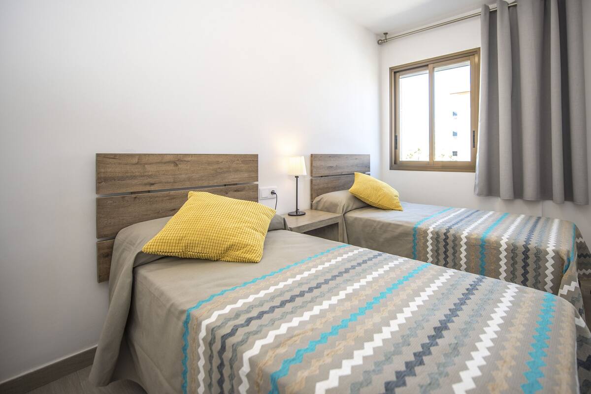 Ein Hotelzimmer mit zwei Betten, jeweils mit einem gemusterten Bettbezug und einem Holz-Kopfteil. Ein Fenster mit Vorhängen und ein Nachttisch mit Lampe befinden sich zwischen den Betten.