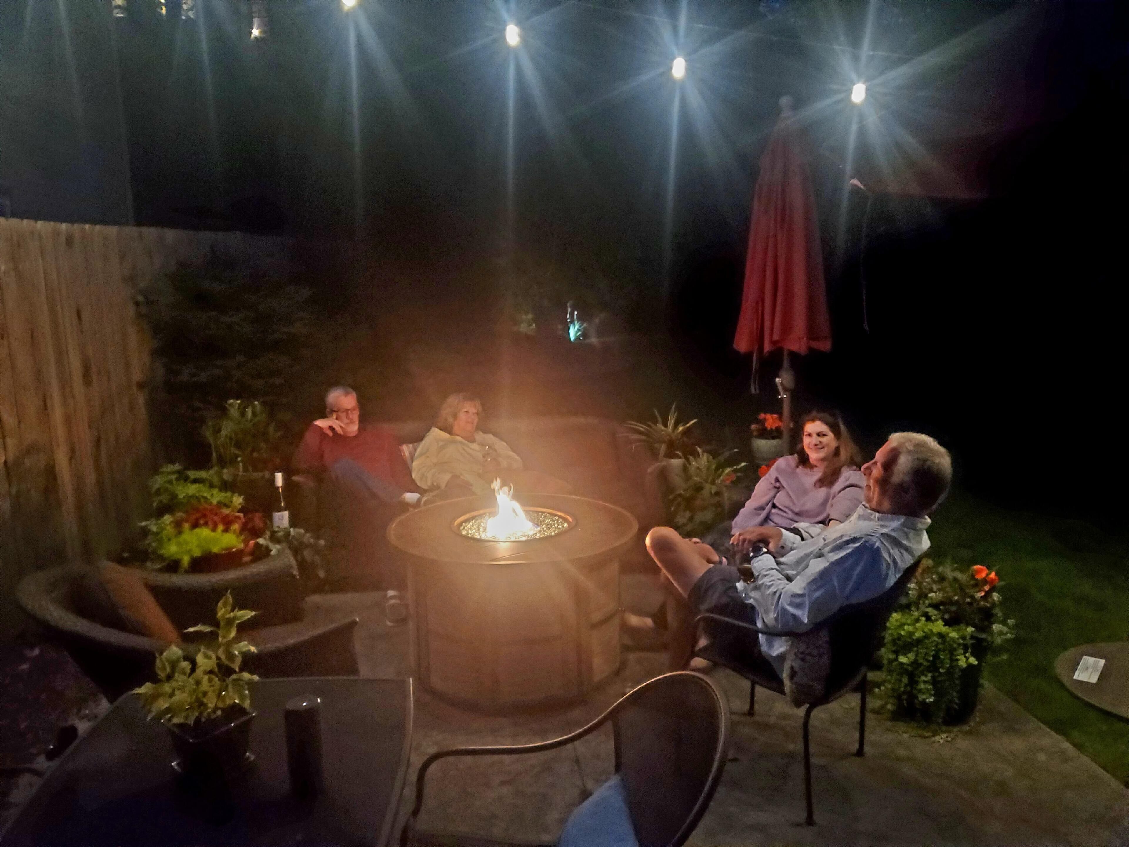 A group of people seated around a circular outdoor fire pit with a table in the center, surrounded by potted plants and outdoor furniture.
