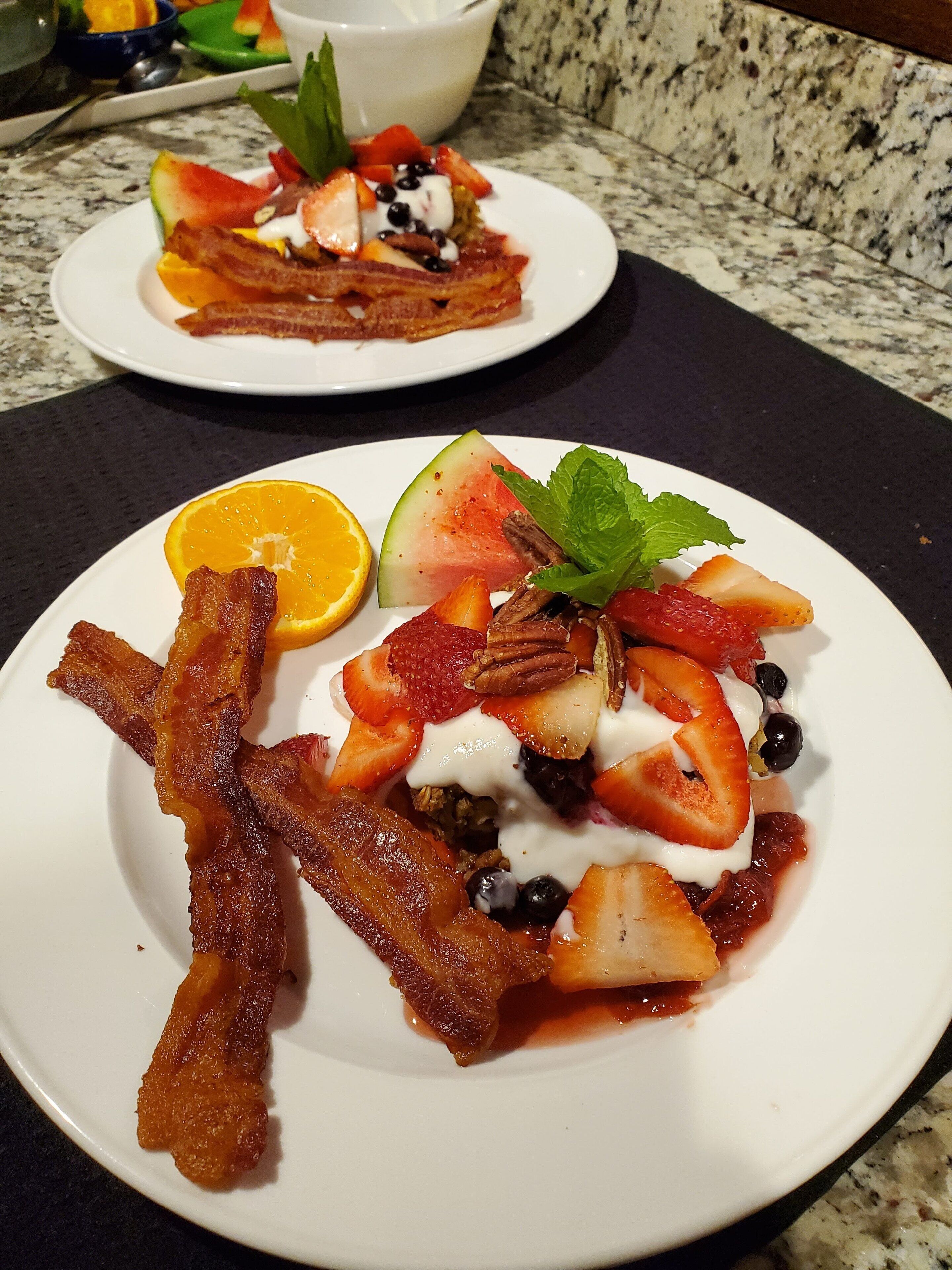 Zwei Teller mit Essen, darunter Speck, Erdbeeren, Blaubeeren und ein Stück Wassermelone auf einer Granitarbeitsplatte.