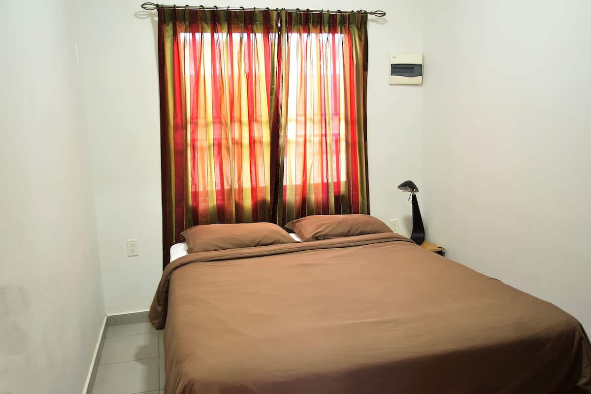A single bed with a brown bedspread and two pillows, a black lamp on a wooden nightstand, and a window with colorful curtains.