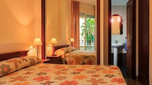 Une chambre d’hôtel comprenant un lit, deux lampes de chevet, une table de chevet, une fenêtre avec des rideaux, une salle de bain avec un lavabo et un miroir, et une vue sur les palmiers à l’extérieur.