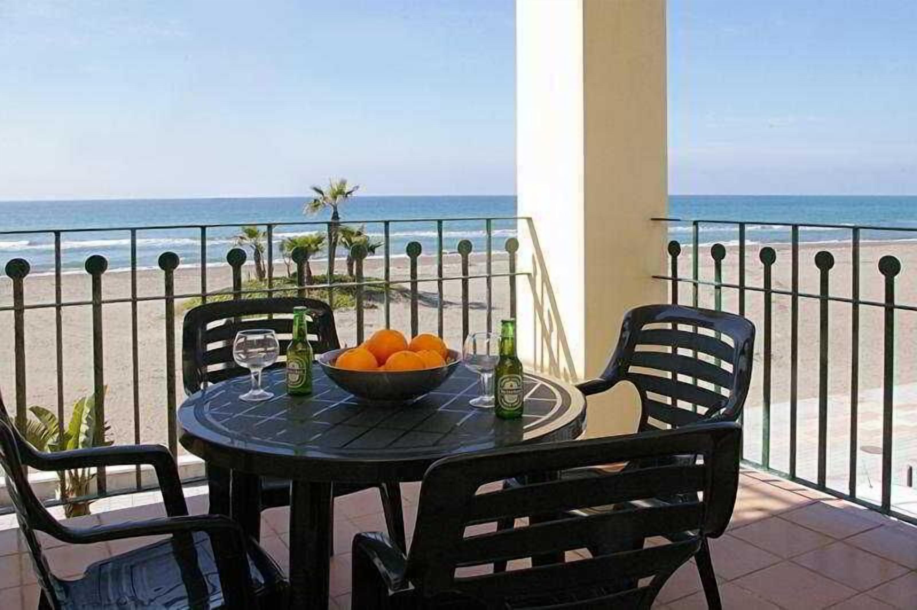 Un balcón con una mesa para dos, que incluye un cuenco de naranjas y dos botellas de cerveza, con vistas a una playa con palmeras.
