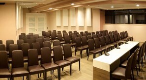Sala de conferência com fileiras de cadeiras e uma longa mesa, equipada com microfones.