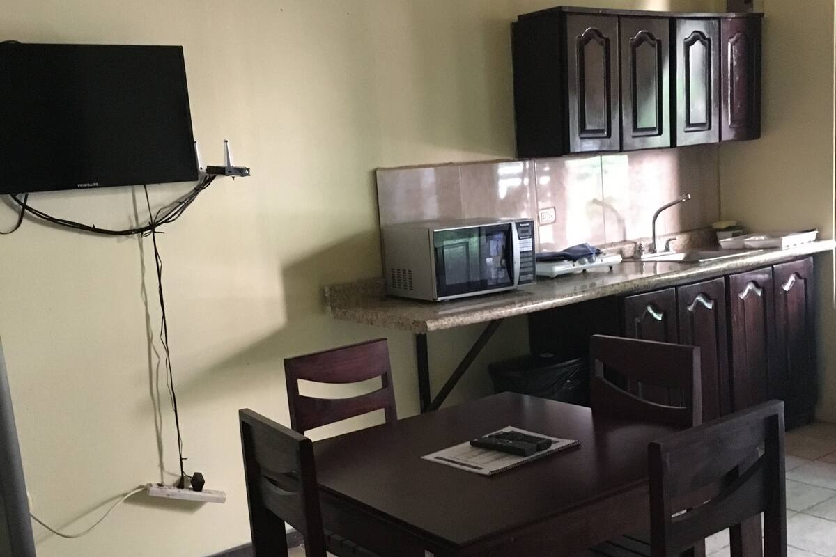 A kitchen with a dining table, chairs, a microwave, cabinets, and a television mounted on the wall.