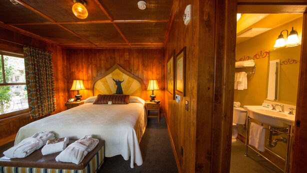 A wooden-themed hotel room with a large bed, two bedside lamps, a headboard with a decorative figure, a small bedside table, and a bench with towels. - McMenamins Old St. Francis (Bend)