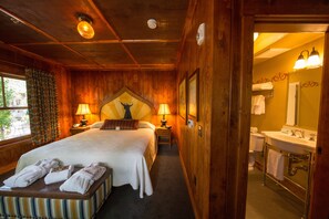A wooden-themed hotel room with a large bed, two bedside lamps, a headboard with a decorative figure, a small bedside table, and a bench with towels. - McMenamins Old St. Francis (Bend)