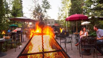 An outdoor seating area with a fire feature, surrounded by tables and chairs, and trees in the background.