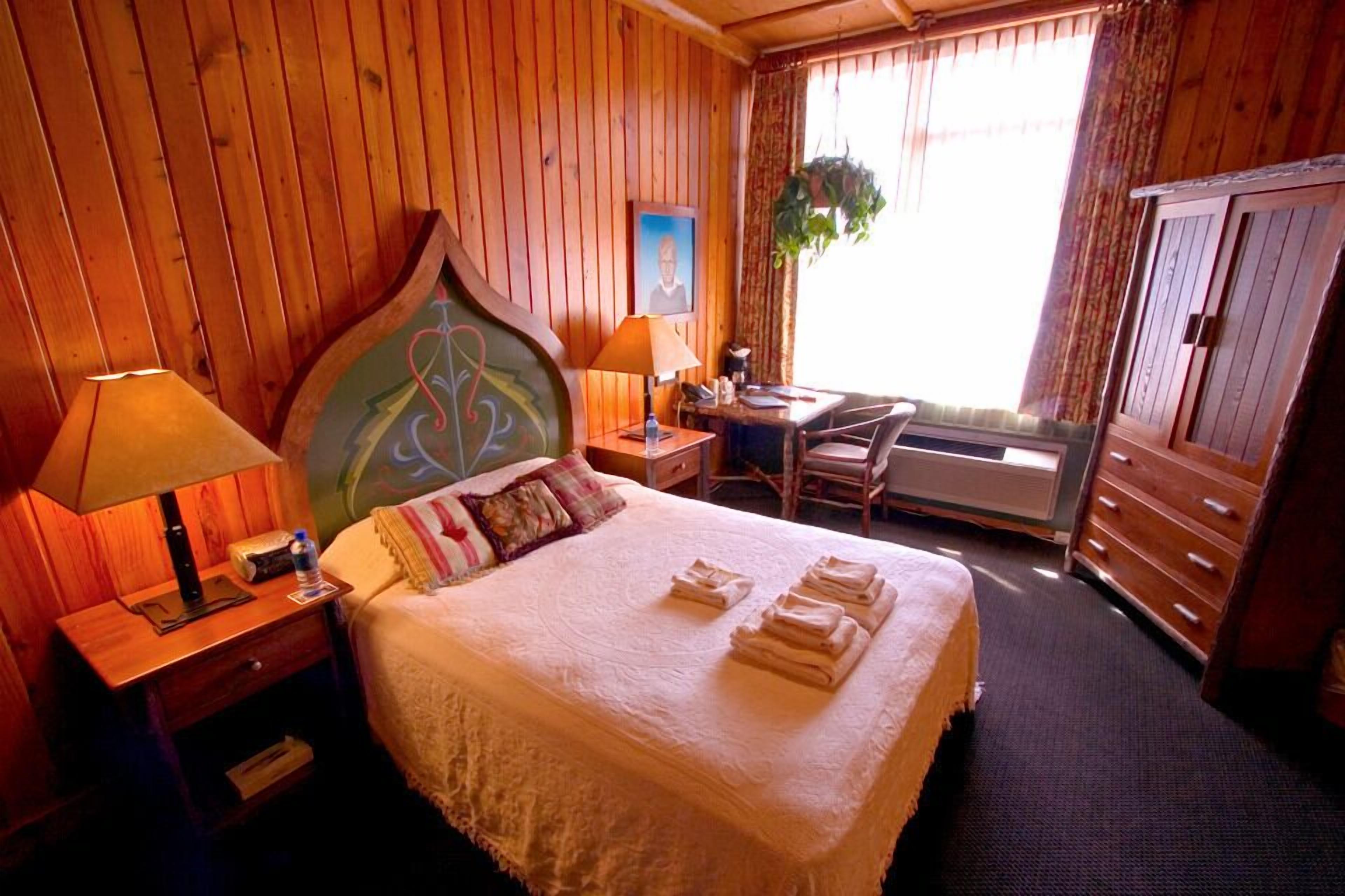 A wooden-paneled room with a bed, two bedside tables, a desk, a chair, and a wardrobe.