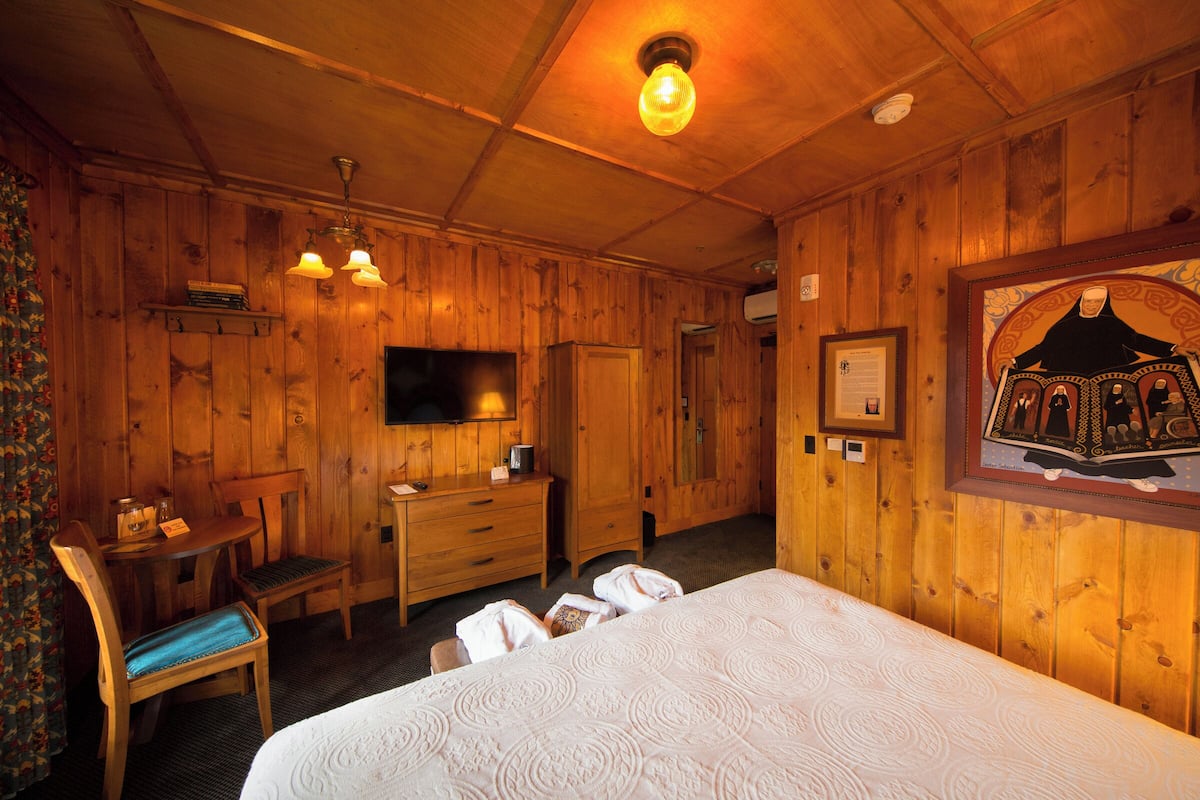 A wooden room with a bed, a desk, a chair, a television, and a framed picture on the wall.