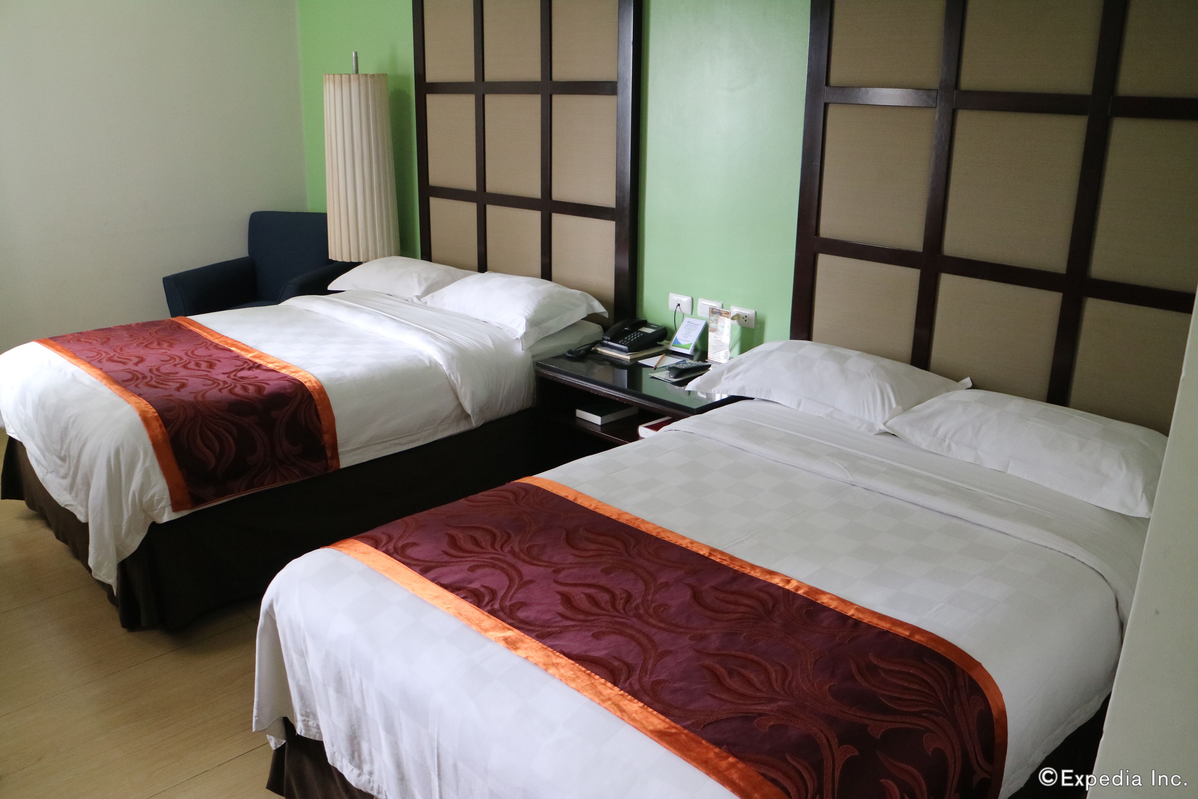 A hotel room with two beds, each with white bedding and a red and brown patterned bedspread. There is a nightstand with a phone, a book, and a tissue box. The room has a green wall and a wooden headboard with a grid pattern.