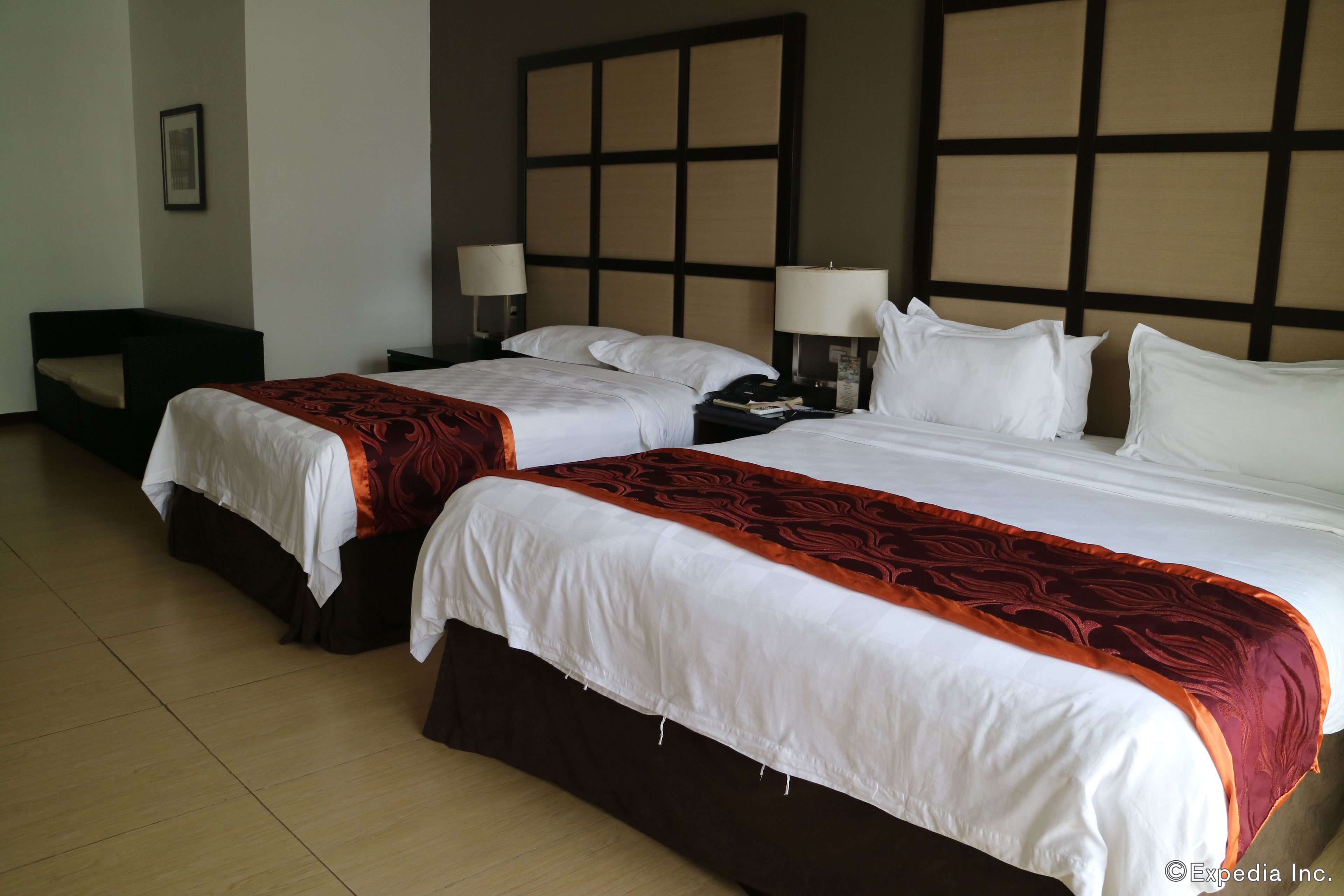 A hotel room with two beds, each with white bedding and a red accent. There are bedside tables with lamps, a large headboard, and a framed picture on the wall.