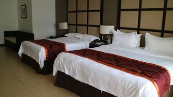 A hotel room with two beds, each with white bedding and a red accent. There are bedside tables with lamps, a large headboard, and a framed picture on the wall.
