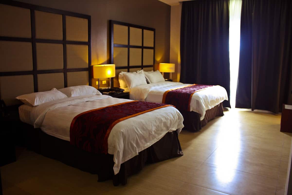 A hotel room with two beds, each with a headboard, bedside tables with lamps, and a large window with curtains.