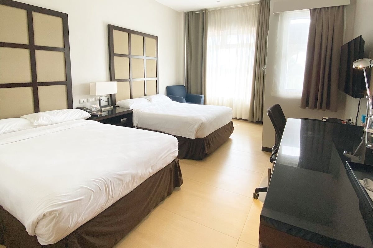 A hotel room with two beds, a desk, a chair, a TV, and a window with curtains.