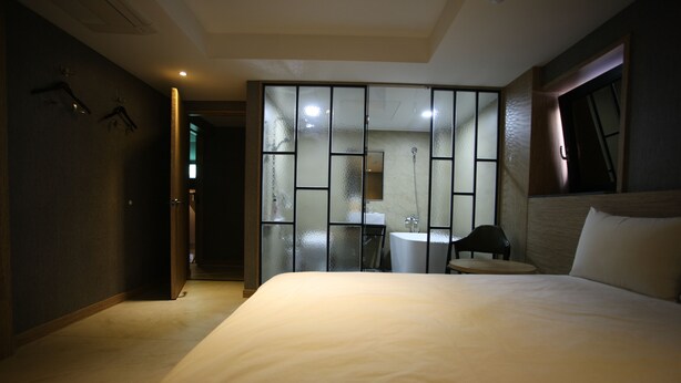 Une chambre d’hôtel avec un grand lit, une petite table, une chaise et une salle de bain visible à travers une cloison en verre dépoli.