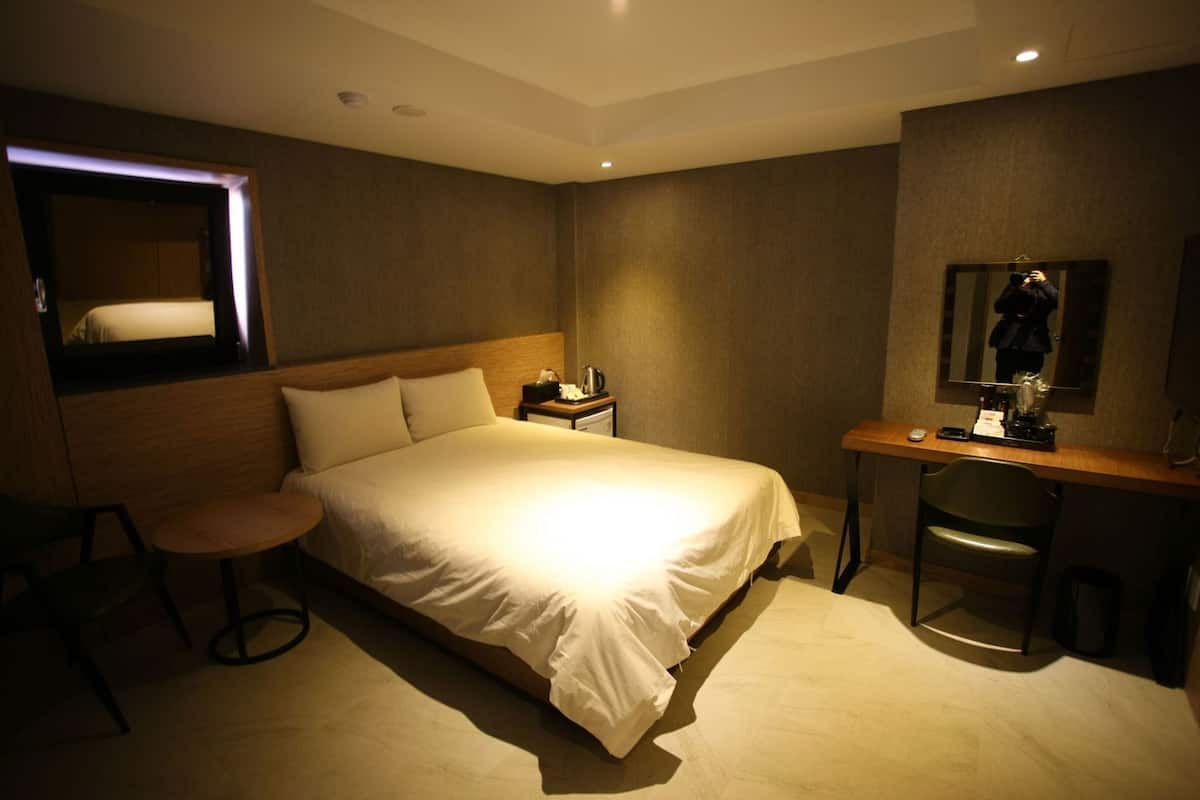 Une chambre d’hôtel avec un lit, un bureau, une chaise, un miroir et un petit tabouret.