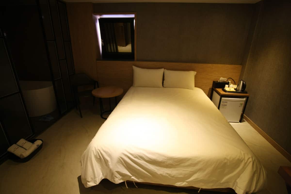 A hotel room with a large bed, a small desk with a lamp, a chair, a nightstand with a phone and a kettle, and a mirror on the wall.