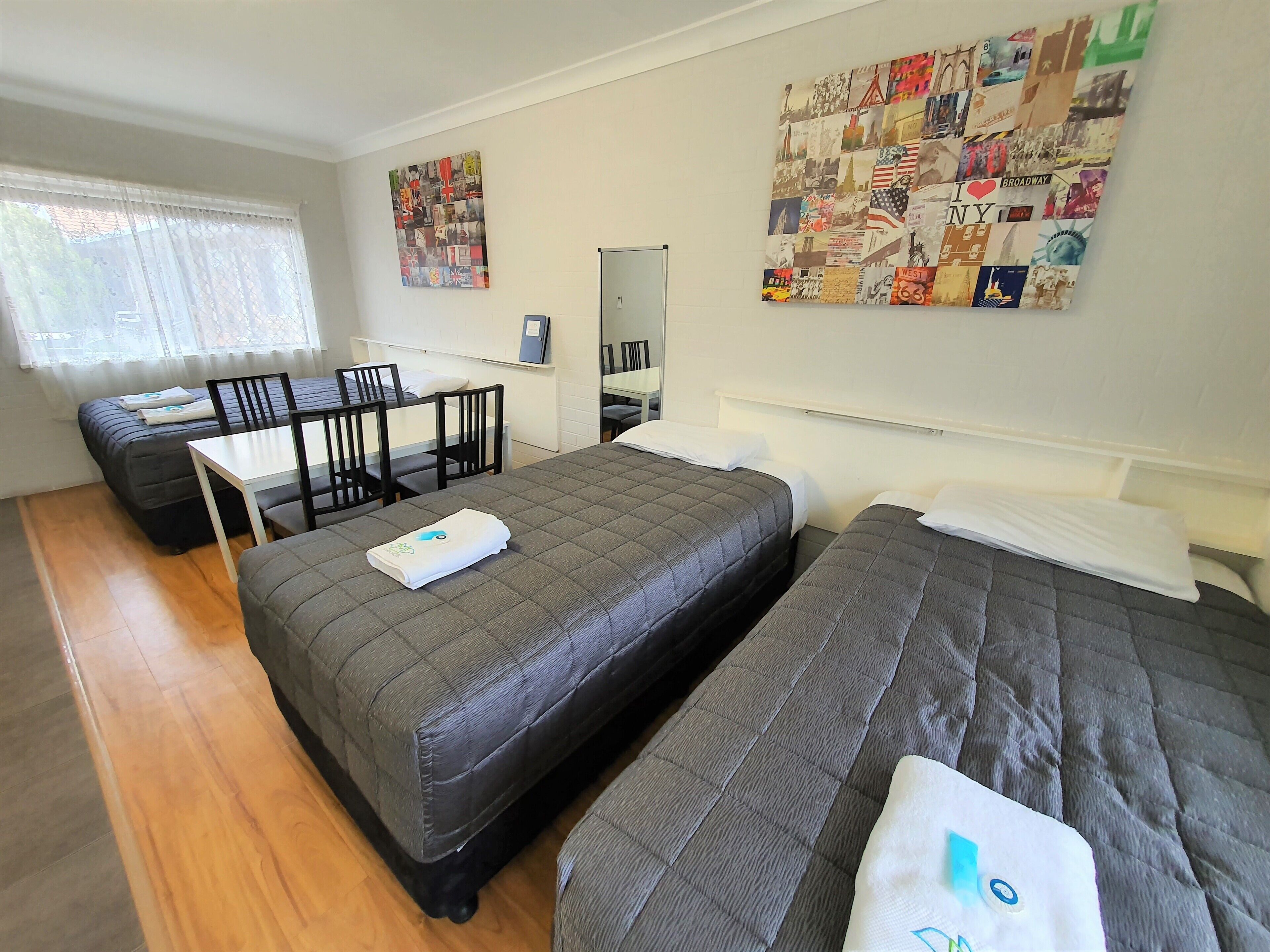 A hotel room with two beds, a desk, and a chair. There is a large collage on the wall and a window with curtains.