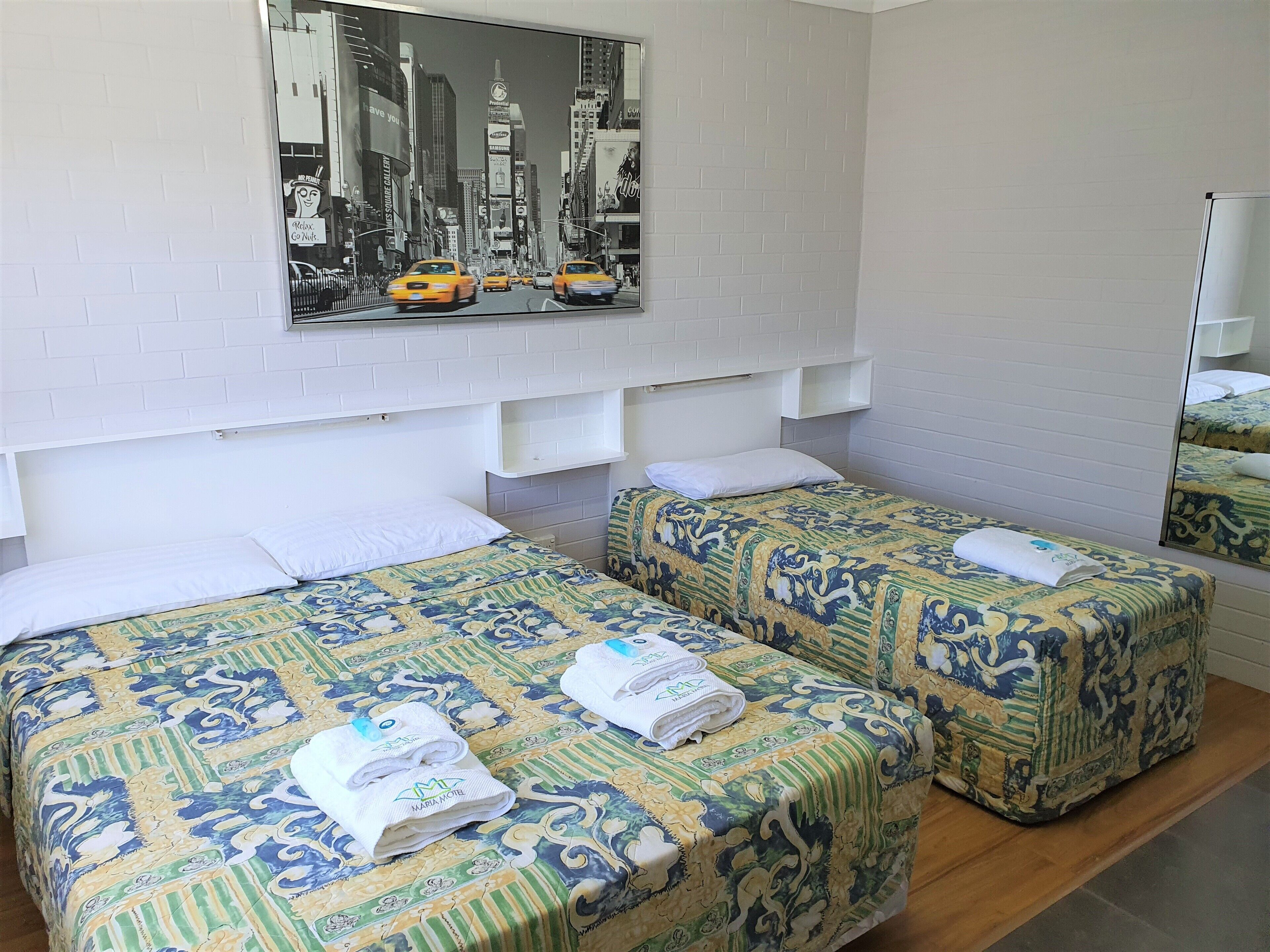 A hotel room with two beds, each with patterned bedding and towels. A large cityscape painting is above the beds, and there is a mirror on the wall.