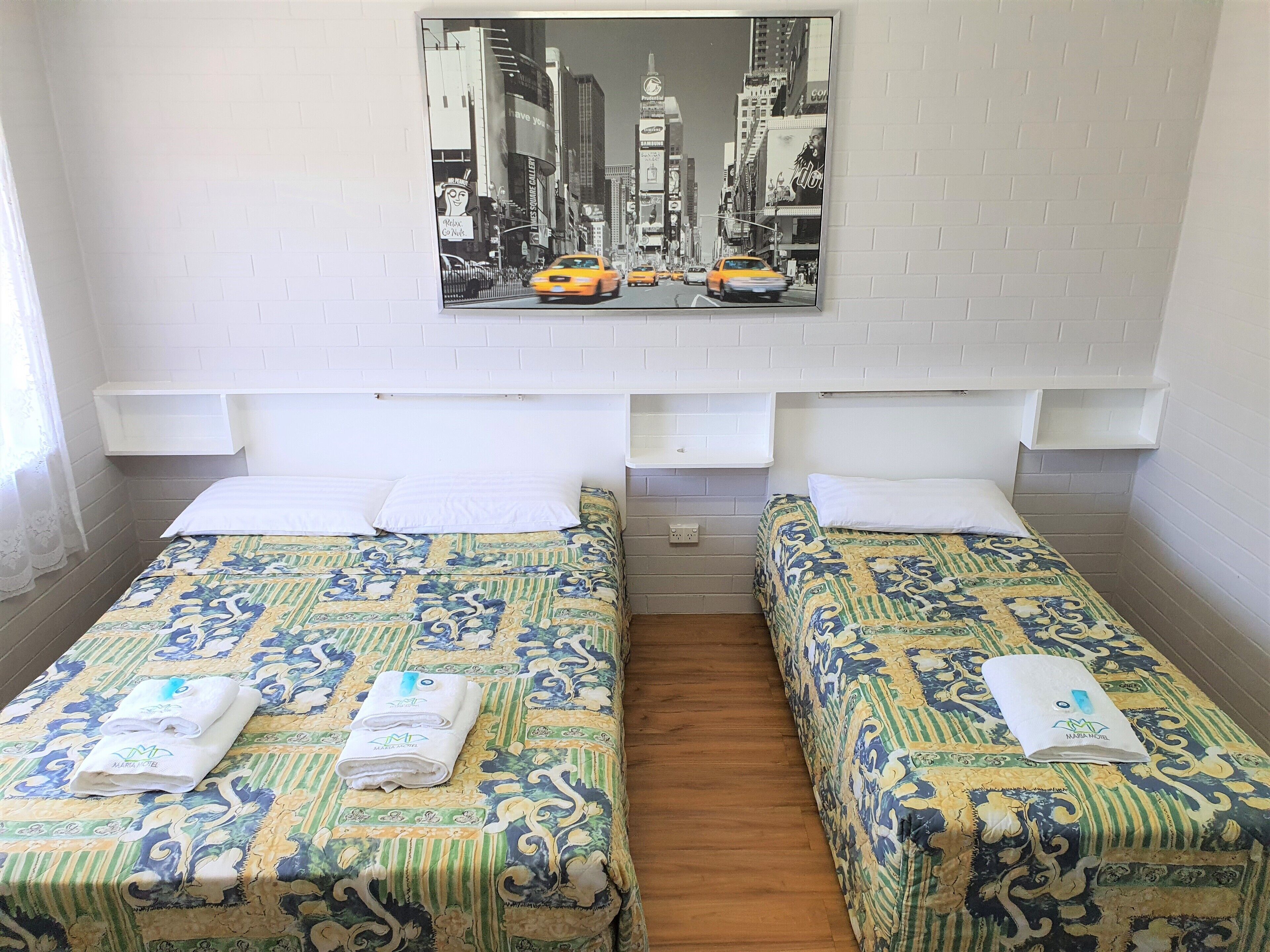 Habitación de hotel con dos camas, cada una con una colcha estampada y una toalla. Se aprecia una pared de ladrillo blanco y una imagen enmarcada de un paisaje urbano sobre las camas.