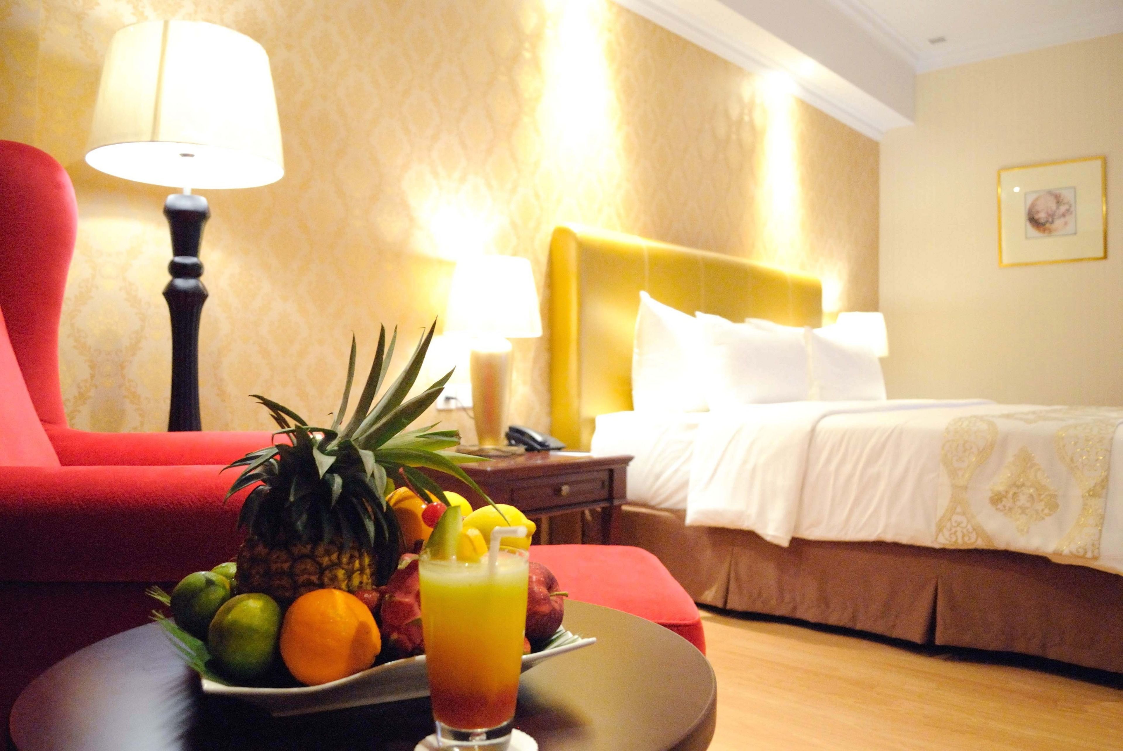 A hotel room with a bed, a red armchair, a nightstand with a lamp, and a table with fruit and a drink.
