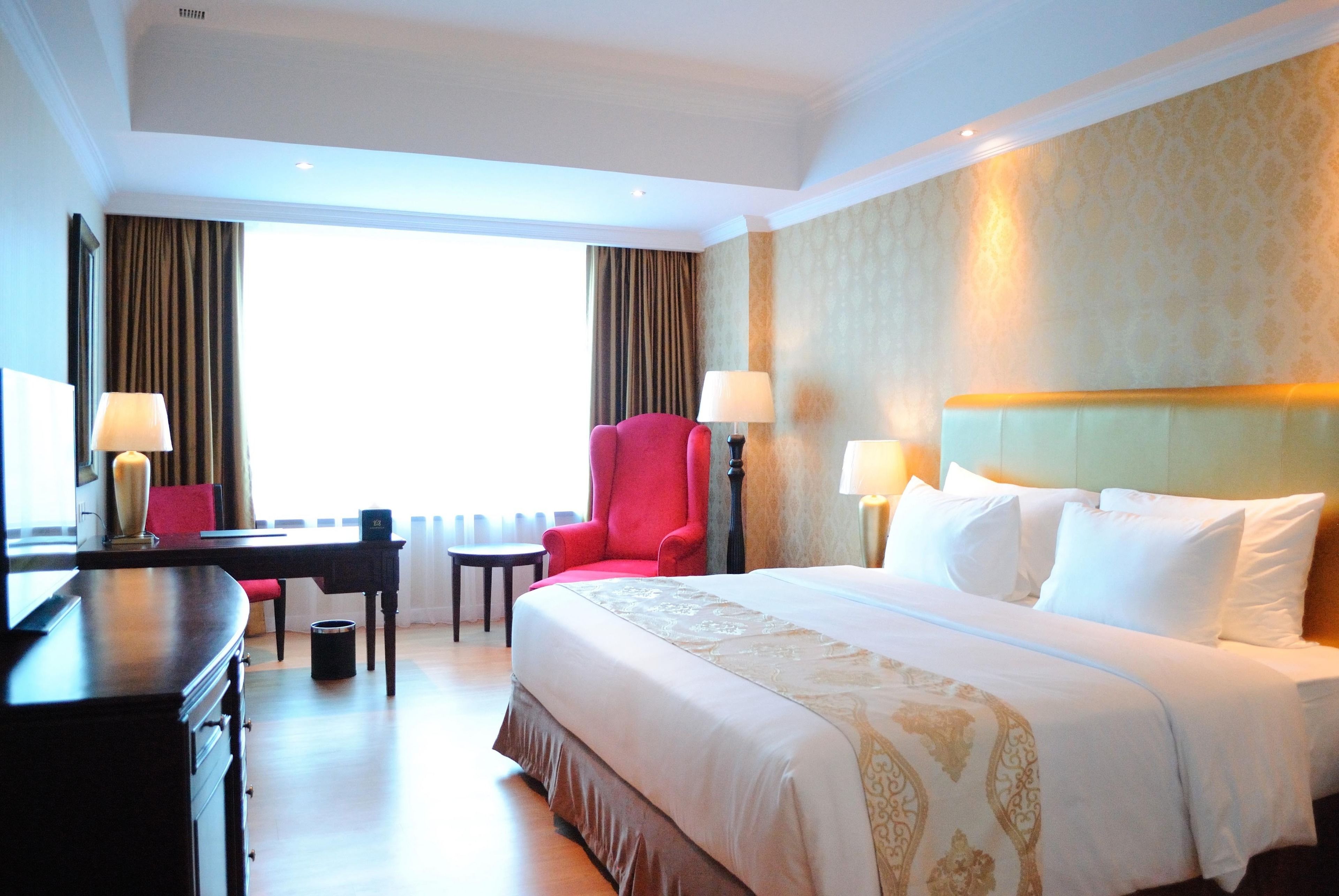 A hotel room with a large bed, a desk, a red armchair, and a window with curtains.