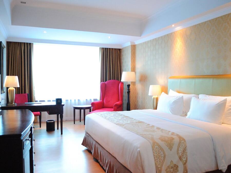 A hotel room with a large bed, a desk, a red armchair, and a window with curtains.