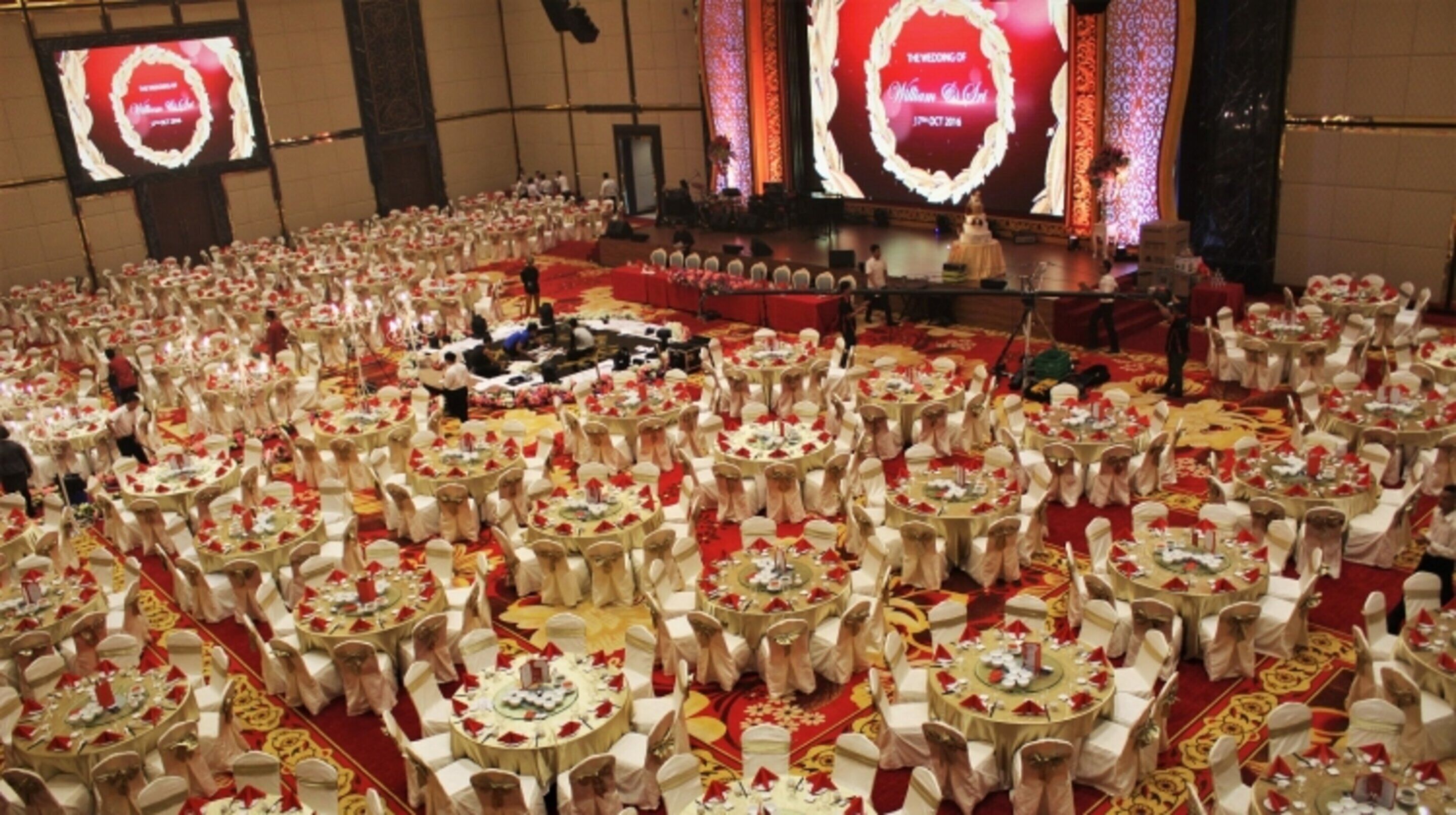 Un gran salón de banquetes con mesas redondas cubiertas con manteles blancos y centros de mesa rojos, dispuestas en un círculo.