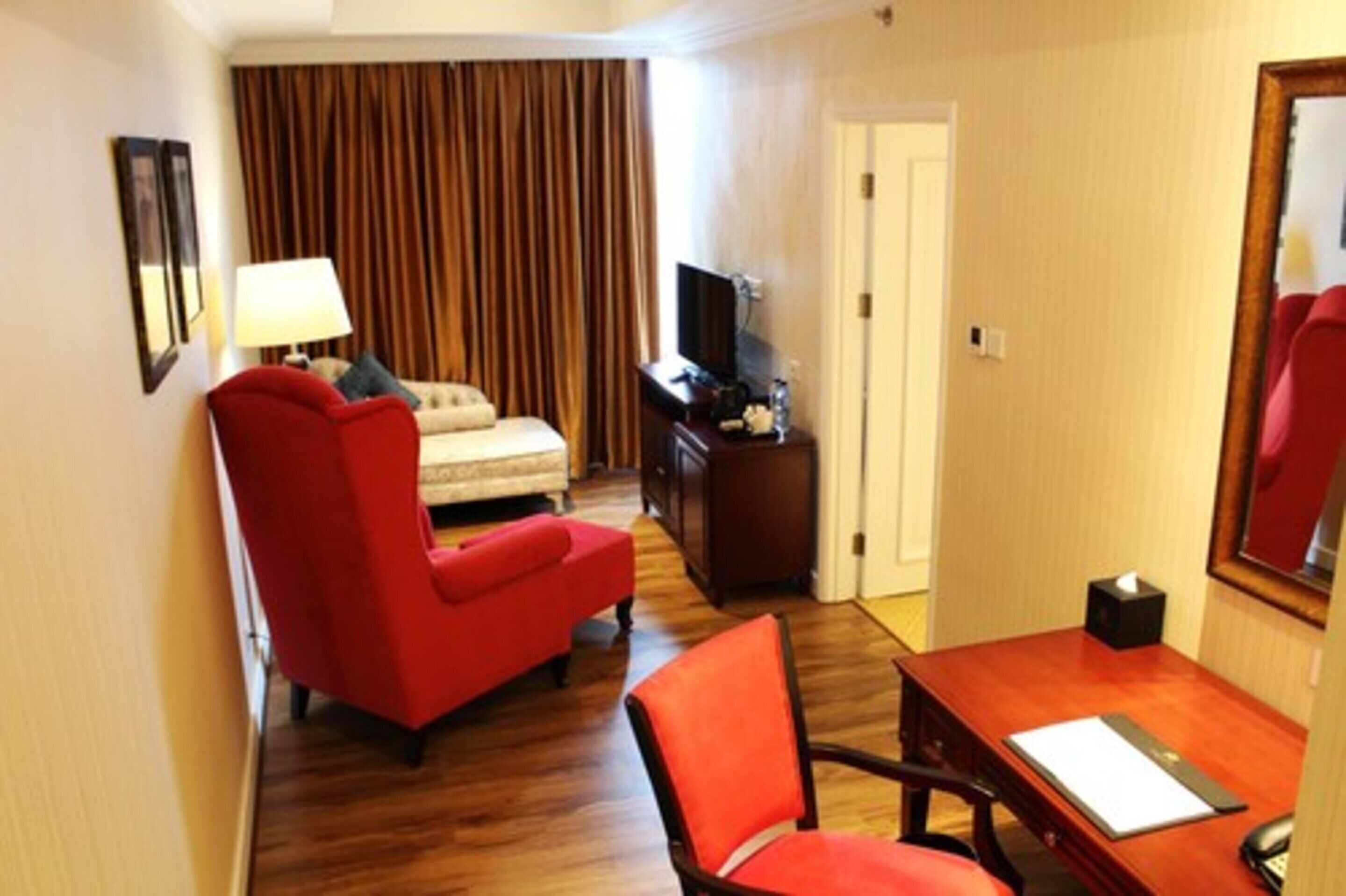 A hotel room with a red armchair, a small sofa, a wooden desk, a television, and a curtain backdrop.