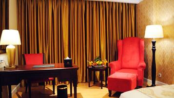 A hotel room with a wooden desk, a red armchair, a small table with fruit and drinks, and two lamps.