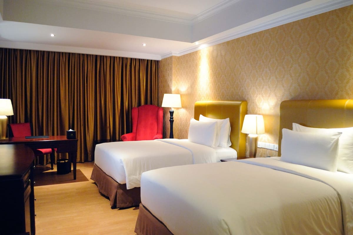 A hotel room with two beds, a desk, a red chair, and a lamp.