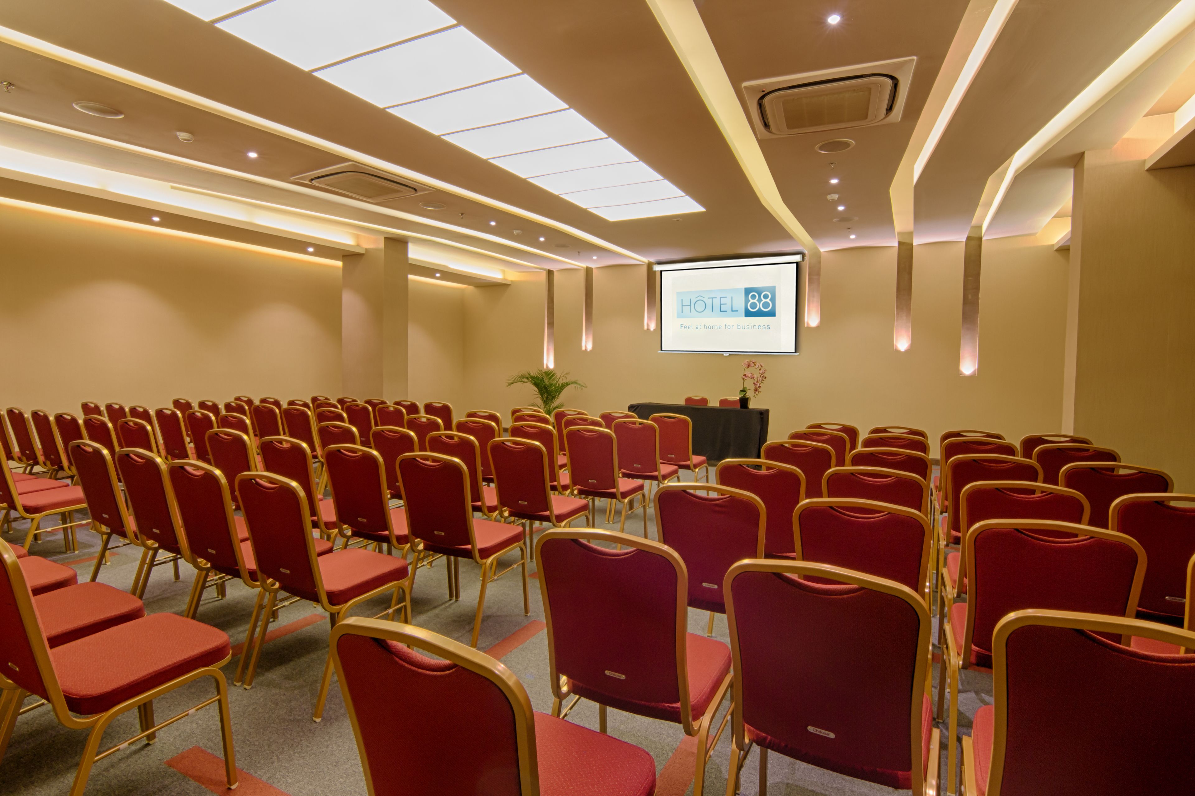 A conference room with rows of red chairs and tables, a projector screen, and a podium.