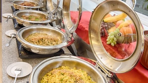 Ein reichhaltiges Buffet mit verschiedenen Speisen in Metallbehältern, eine Obstauslage und ein Fenster mit Jalousien.