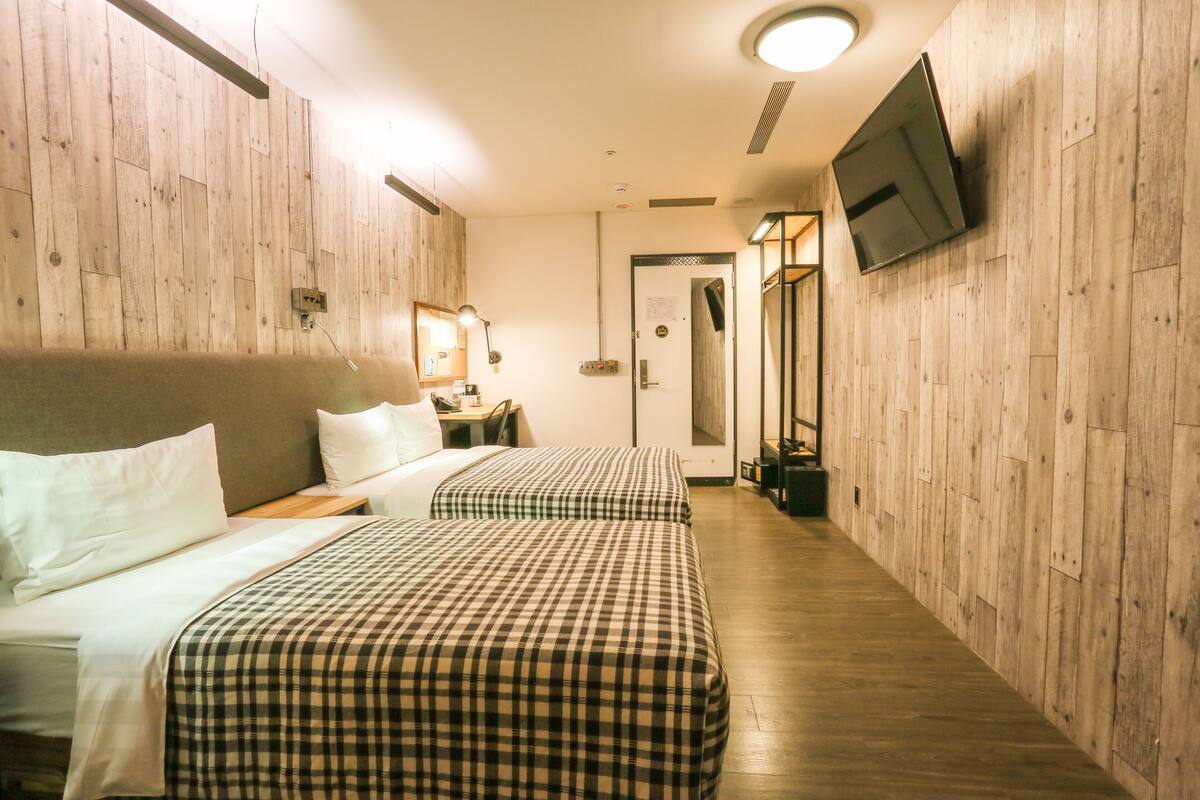 A hotel room with two beds, wooden walls, a TV mounted on the wall, and a small desk with a lamp.