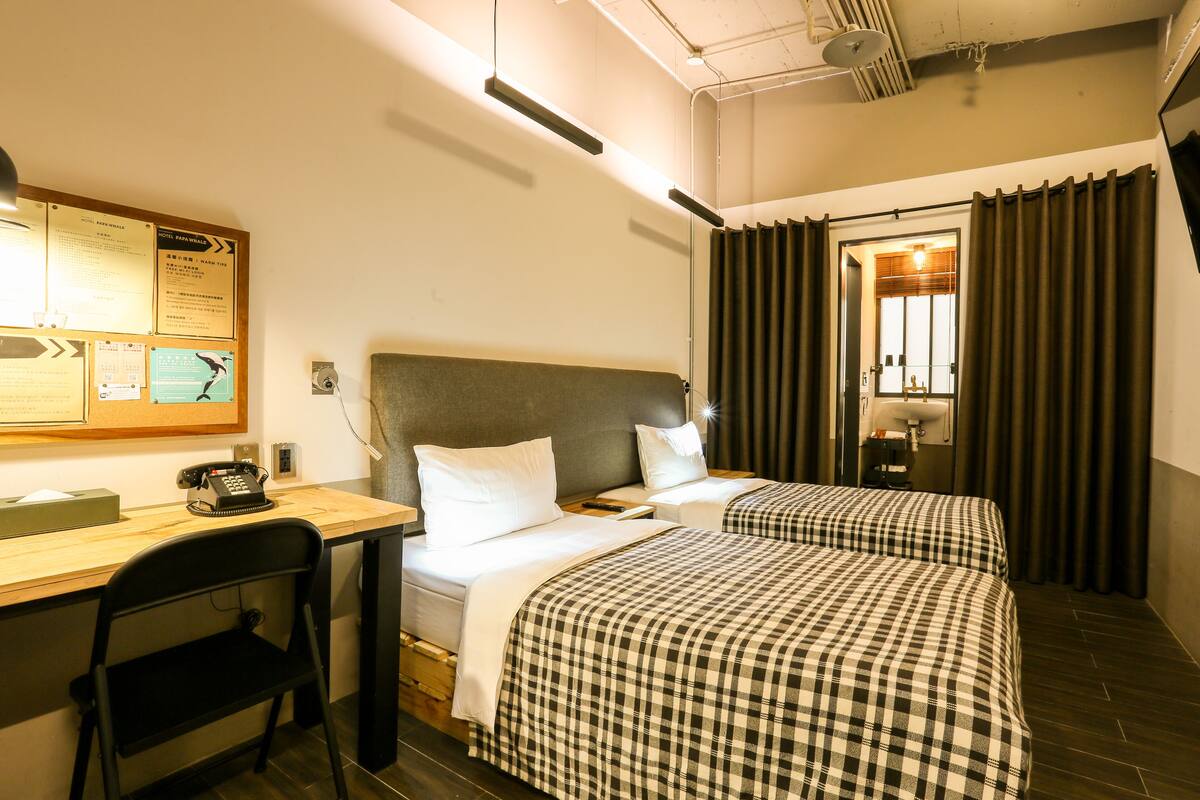 A hotel room with two beds, a desk, a chair, a bulletin board, and a bathroom visible through a door.