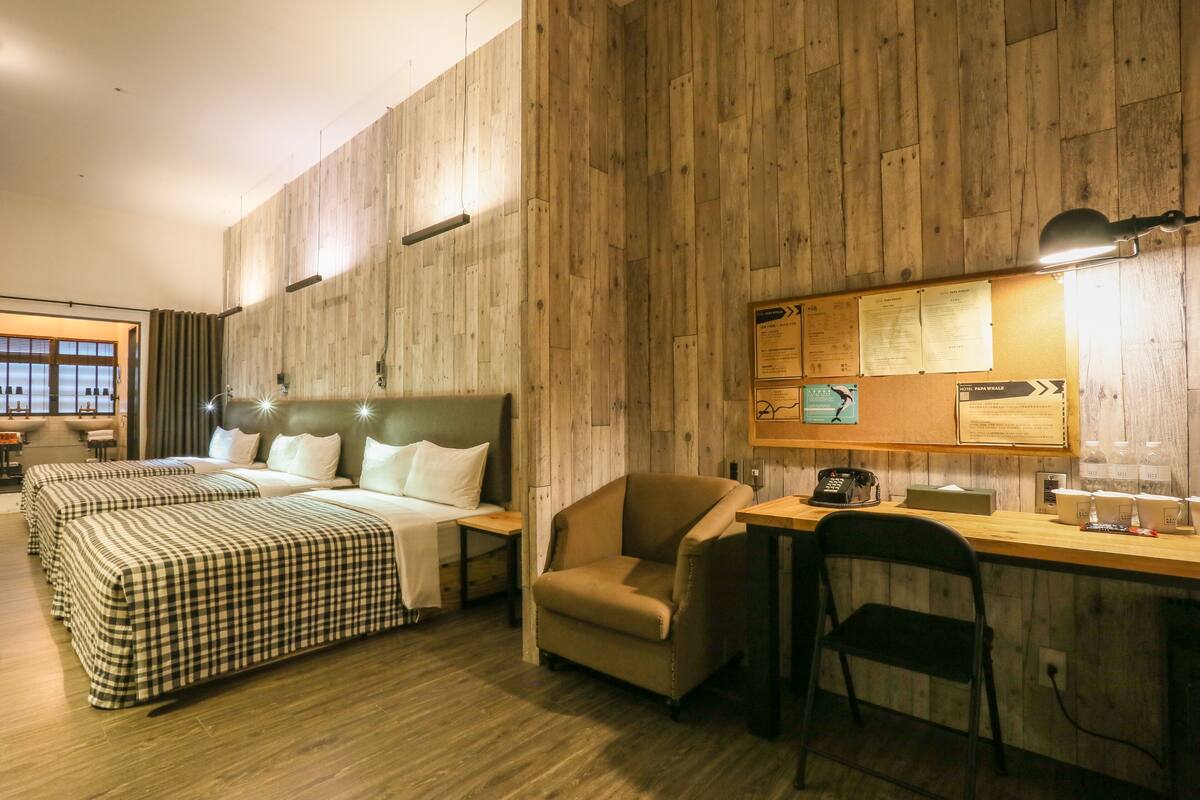 A hotel room with a wooden wall, a bed with checkered bedding, a desk with a lamp, and a chair.