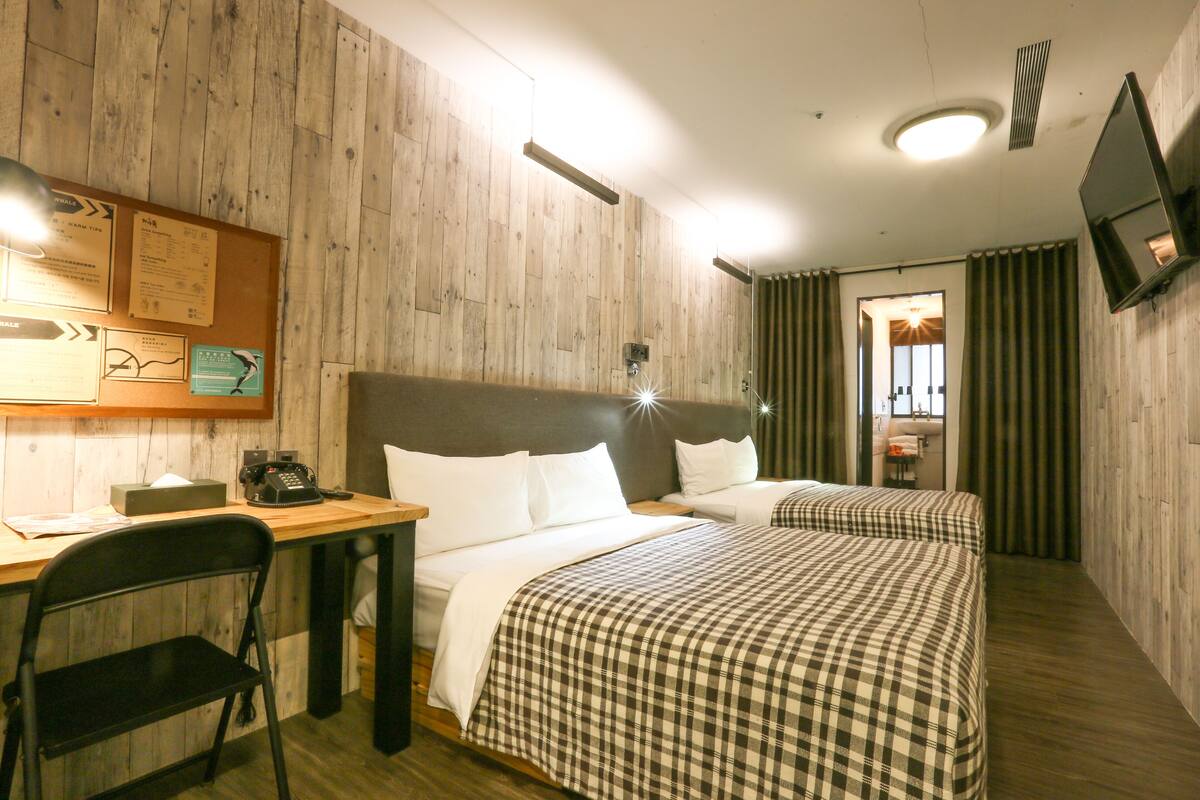 A hotel room with two beds, a desk, a chair, a bulletin board, and a TV mounted on the wall.