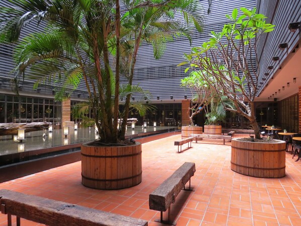 An indoor atrium with a high ceiling, large windows, and a central water feature. There are wooden planters with trees and benches around the water.