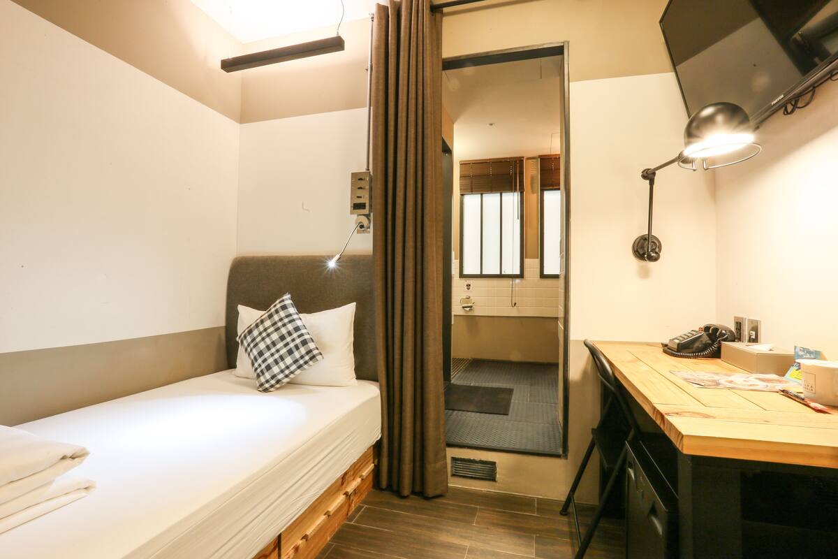 A compact hotel room with a single bed, a desk, a chair, a desk lamp, and a television.