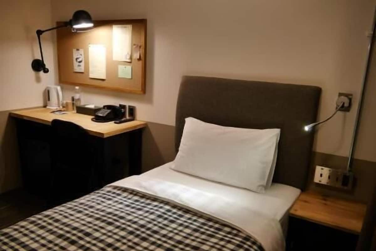 A hotel room with a bed, a desk, a chair, a lamp, and a bulletin board.