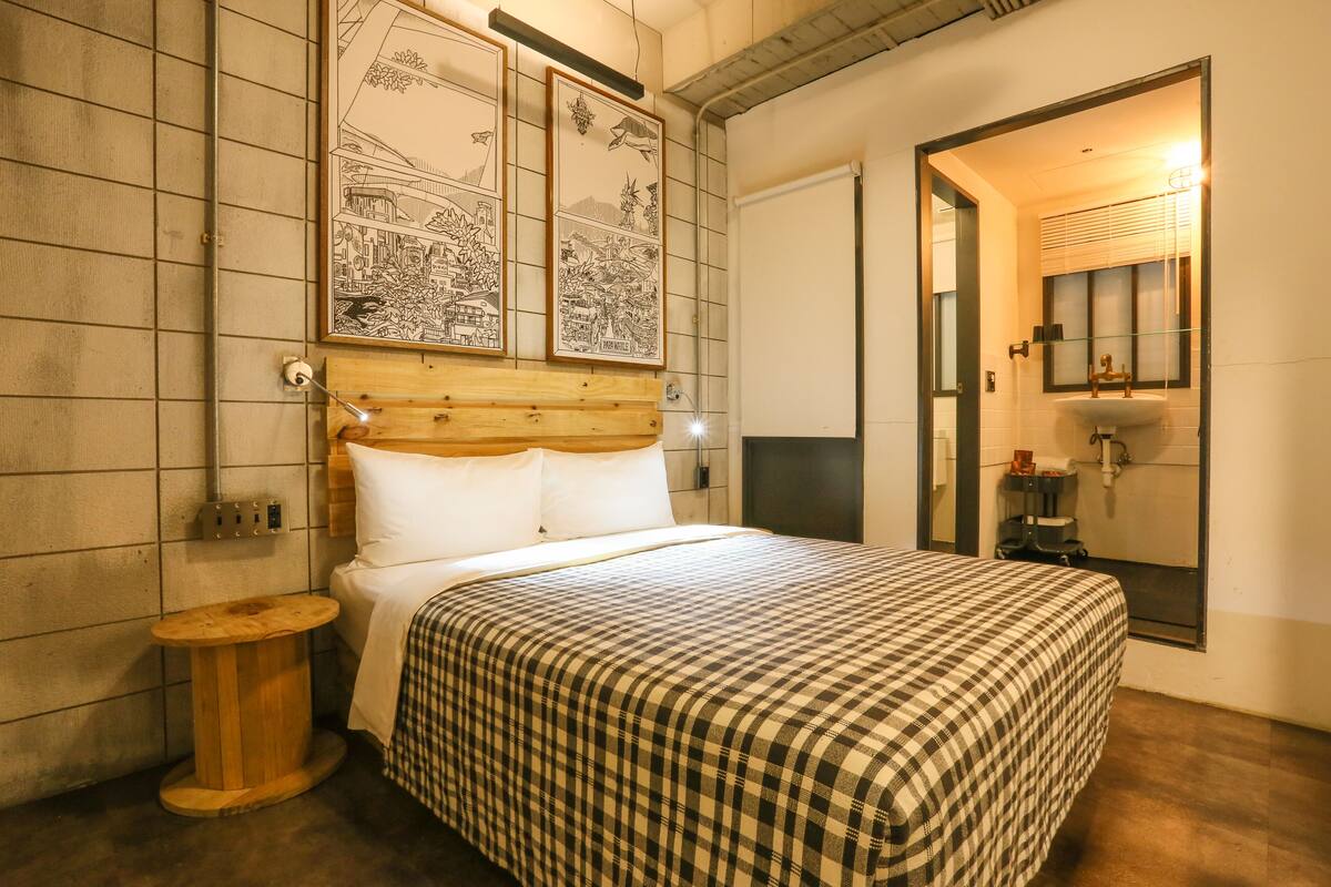 A neatly made bed with a checkered bedspread, a wooden headboard, and a small wooden stool beside it.