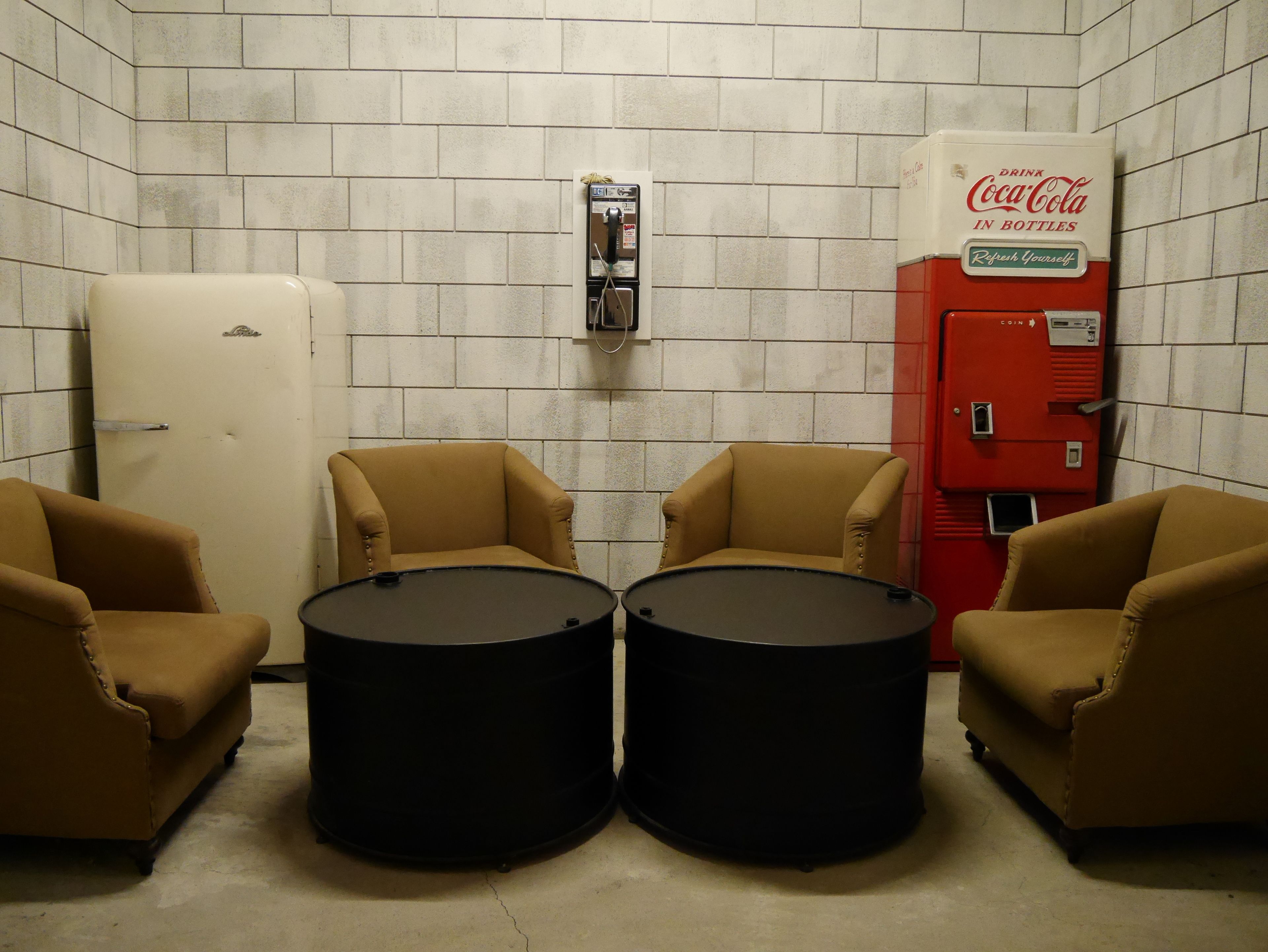 A room with two armchairs, a white refrigerator, a red Coca-Cola vending machine, and two black barrels.