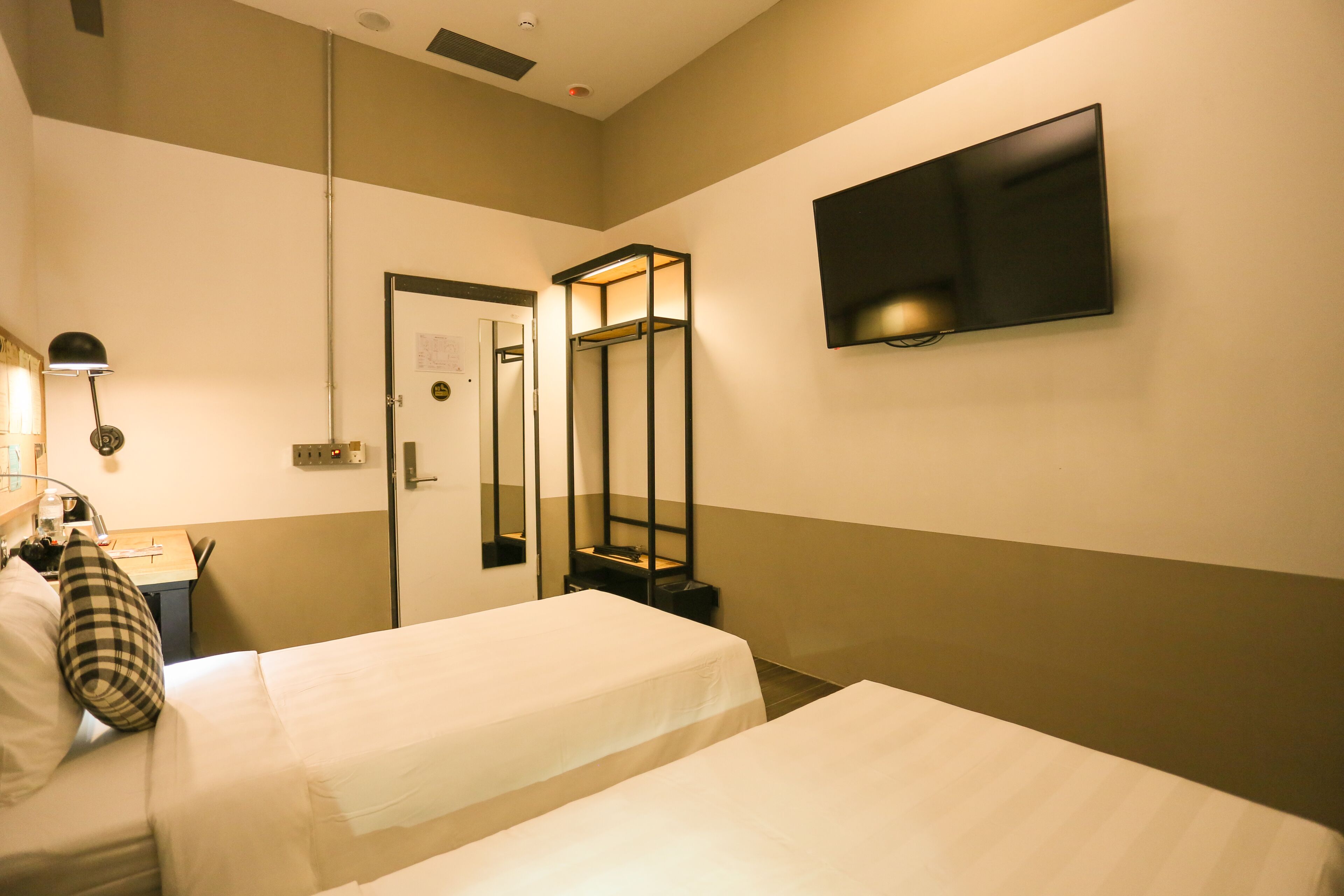 A hotel room with two single beds, a desk with a chair, a flat-screen TV mounted on the wall, and a mirror with a shelf.